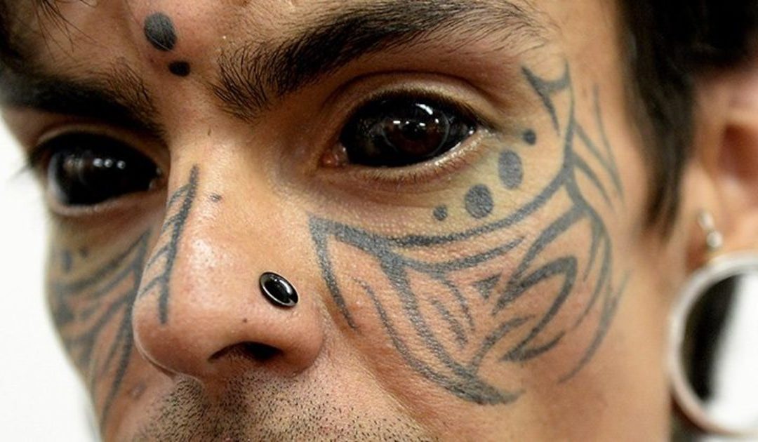 a face of a person whose skin is covered in tattoos, and their eye balls have been permanently tattooed black.