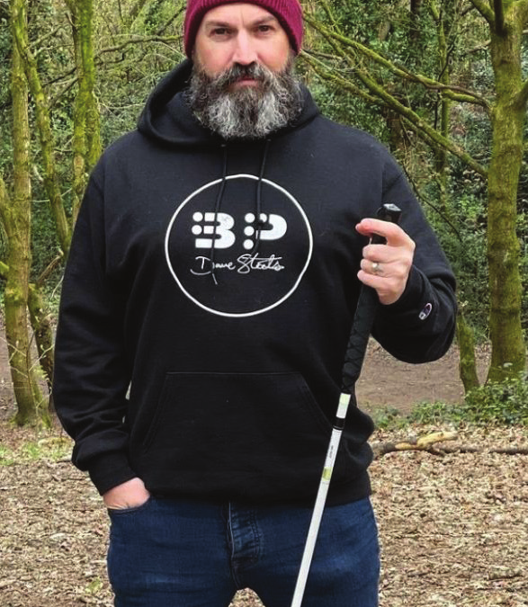 Poet David Steele holds a white cane and stands outside in a black sweatshirt, jeans, and a red hat.