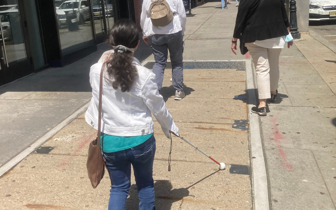 A VLANJ program participant holds a white cane and walks the streets of Morristown behind two individuals.