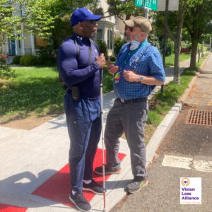 Two individuals stand at a street corner in Morristown. One holds a white cane.